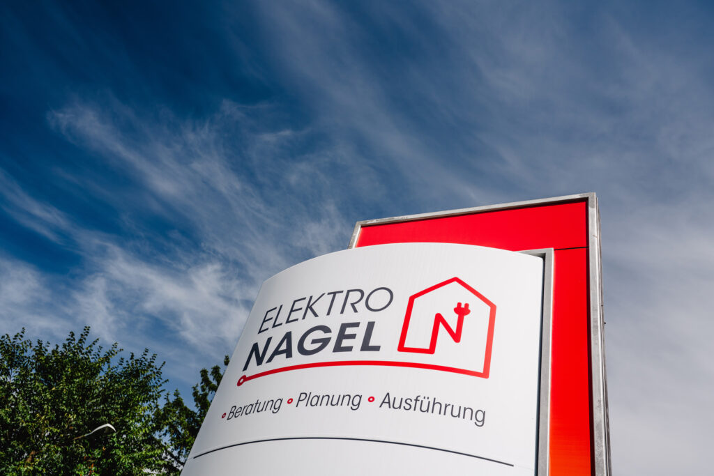 Ein Hinweisschild mit dem Firmenlogo "Elektro Nagel" sowie dem Text "Beratung, Planung, Ausführung". Im Hintergrund ist blauer Himmel mit leichter Bewölkung zu sehen.
